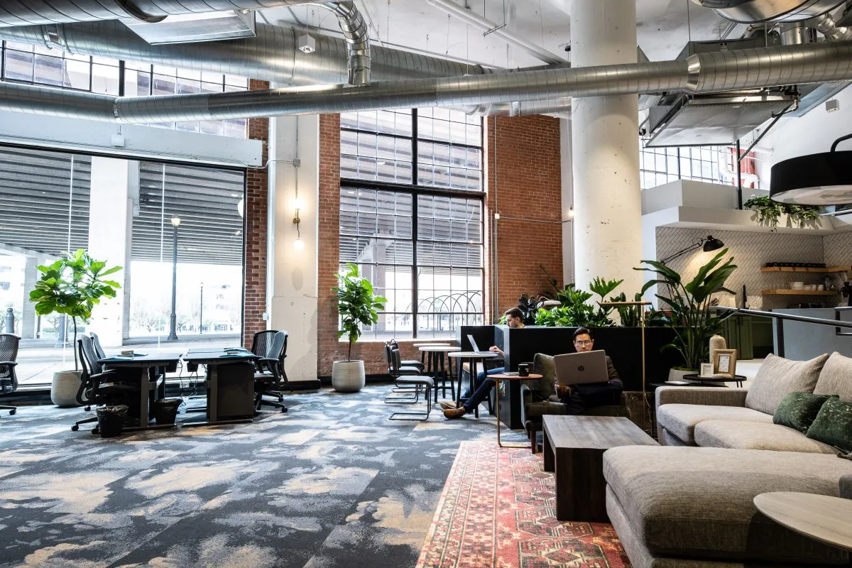 Common Desk at 6th and Congress with exposed brick, industrial ductwork, large windows, lounge seating, and coworkers on laptops
