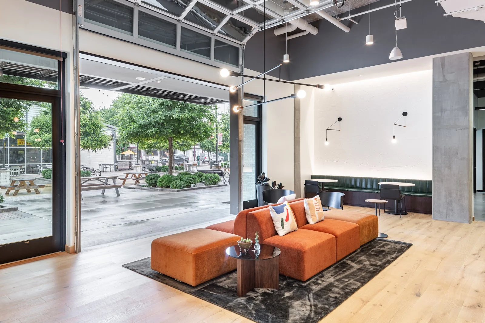 Industrious Pearl Expressway lounge with large roll-up garage door opening to a tree-lined courtyard with picnic tables, burnt orange modular sectional sofa on dark rug, dark green leather banquette with round white tables, concrete column, swing-arm wall lights, and warm oak floors