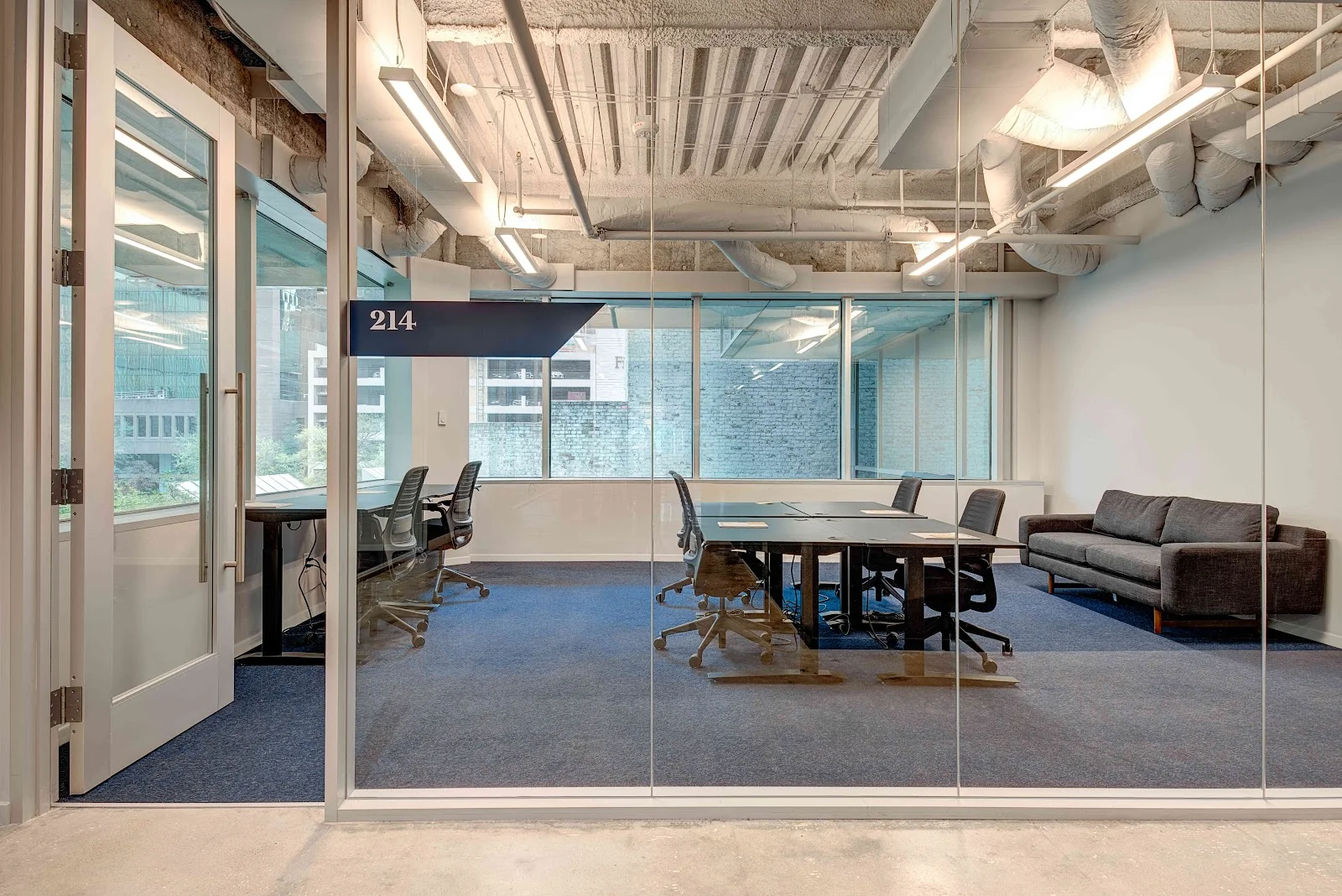 The Cue Arts District glass-walled conference room labeled 214 with blue carpet, dark conference table surrounded by ergonomic mesh chairs, grey sofa along the wall, exposed concrete ceiling with linear LED fixtures and wrapped ductwork, and city-view windows
