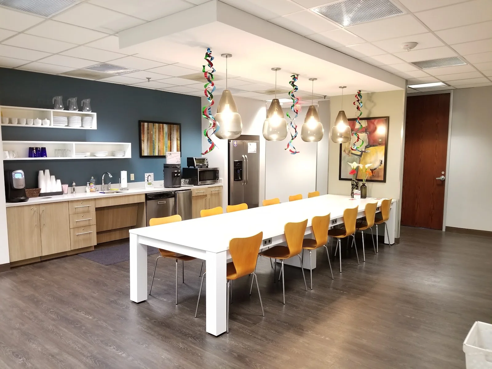 HQ Highland Park Place communal kitchen with long white table surrounded by amber bentwood chairs, teal accent wall with open shelving displaying white dishes, teardrop pendant lights with colorful ribbon streamers, stainless appliances, and grey vinyl plank floors