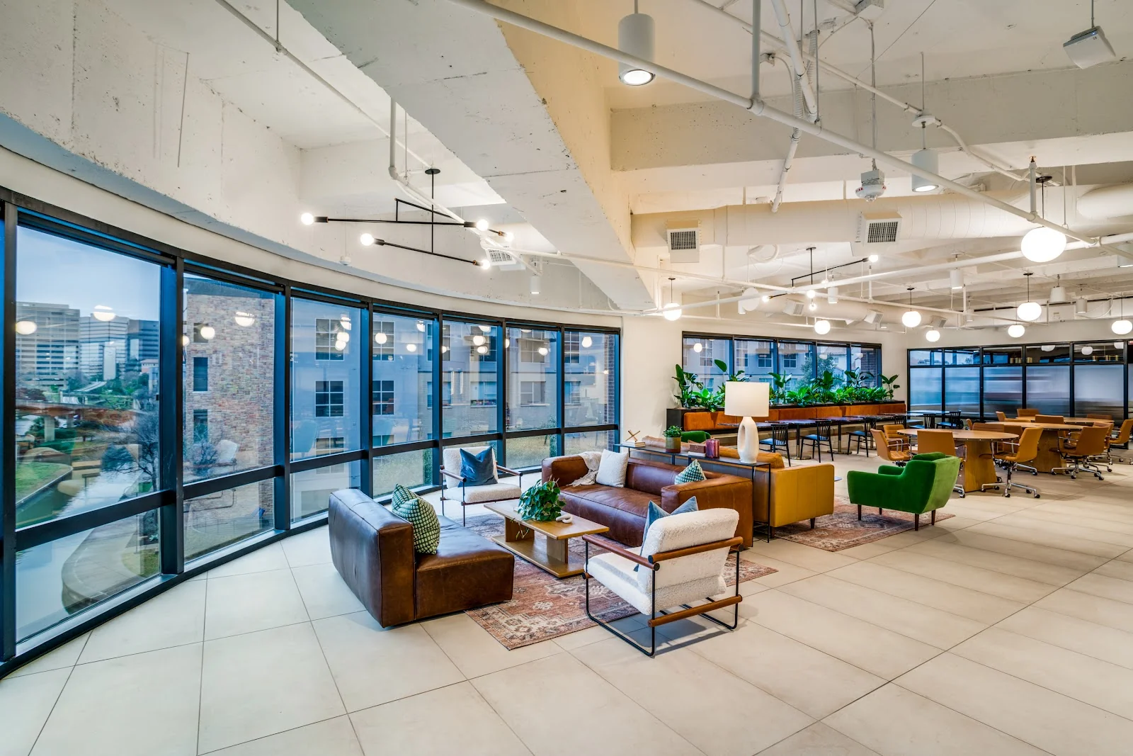 Foster Coworking Canal Centre luxurious curved-glass lounge with floor-to-ceiling black-framed windows overlooking a canal at dusk, brown leather sofas and cream armchairs on vintage Persian rug, green wingback chair, exposed white ceiling with industrial ductwork, globe lights, and plant dividers
