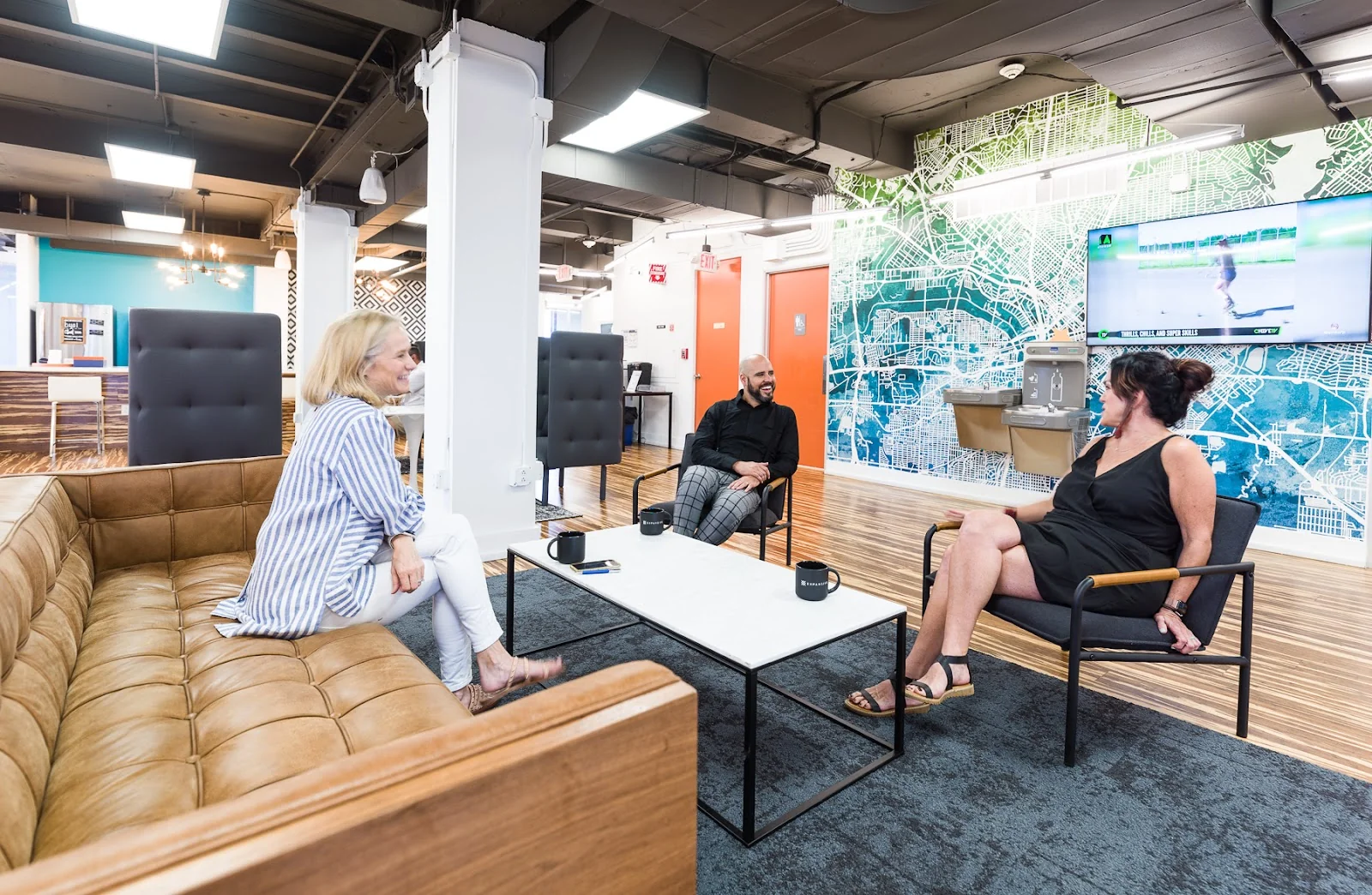 Expansive Katy Building vibrant lounge with tan tufted leather sectional, white marble-top coffee table, black modern armchairs, large teal city-map wall mural, wall-mounted TV, exposed black ceiling, warm hardwood floors, and three members chatting casually
