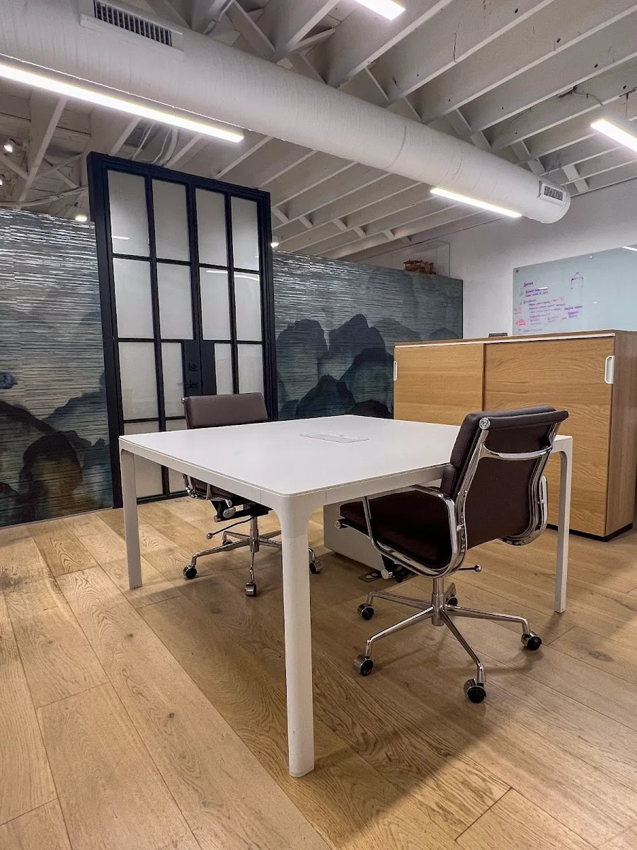 Spryrocket Station private office with warm oak hardwood floors, white desk with two brown leather Eames-style chairs, dramatic blue-grey mountain landscape wallpaper mural, black-framed frosted glass partition door, oak storage cabinet, whiteboard, and exposed white ceiling with linear LED fixtures