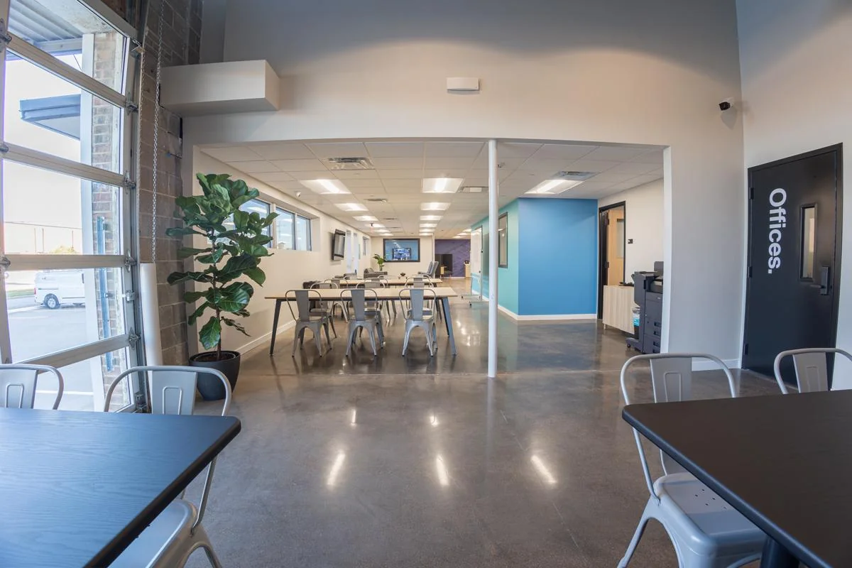 Polygon converted industrial space with roll-up garage door windows flooding natural light, polished concrete floors, silver Tolix-style metal chairs at dark communal tables, fiddle-leaf fig plant, teal accent wall, Offices sign on dark door, and clerestory windows