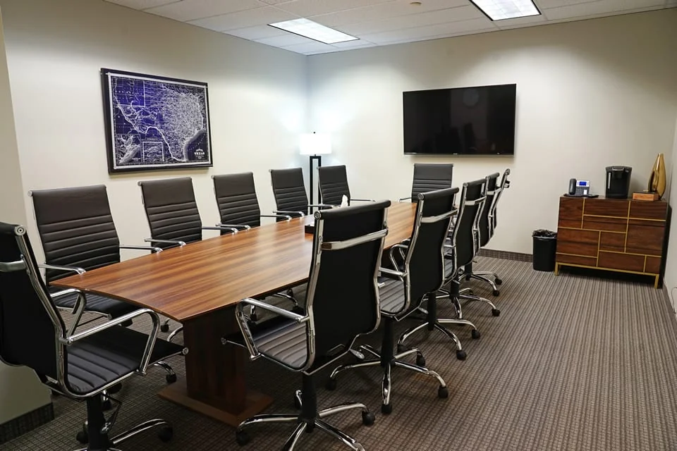 ExecutiveWorkspace Uptown Dallas traditional boardroom with long walnut conference table, black leather high-back executive chairs, framed vintage Texas map on wall, wall-mounted flat-screen TV, walnut credenza with Keurig, and striped commercial carpet