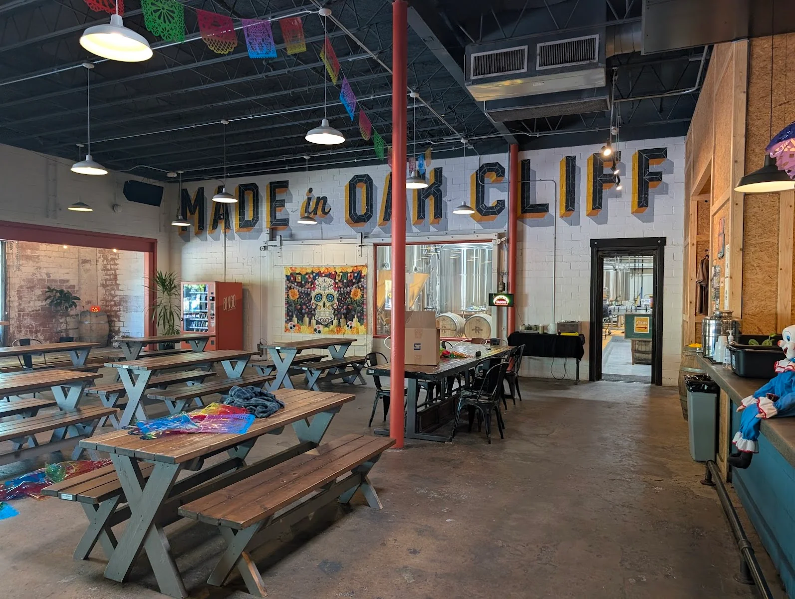 Tyler Station brewery-style taproom with Made in Oak Cliff painted in bold block letters on white cinder block wall, wooden picnic tables, red support columns, colorful papel picado banners, sugar skull artwork, and brewing equipment visible through doorway