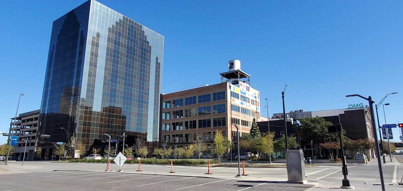 2201 Main exterior street view of downtown Dallas showing dark glass office tower and adjacent four-story brown brick building with colorful rooftop art installation and water tower, clear blue sky, and tree-lined sidewalks