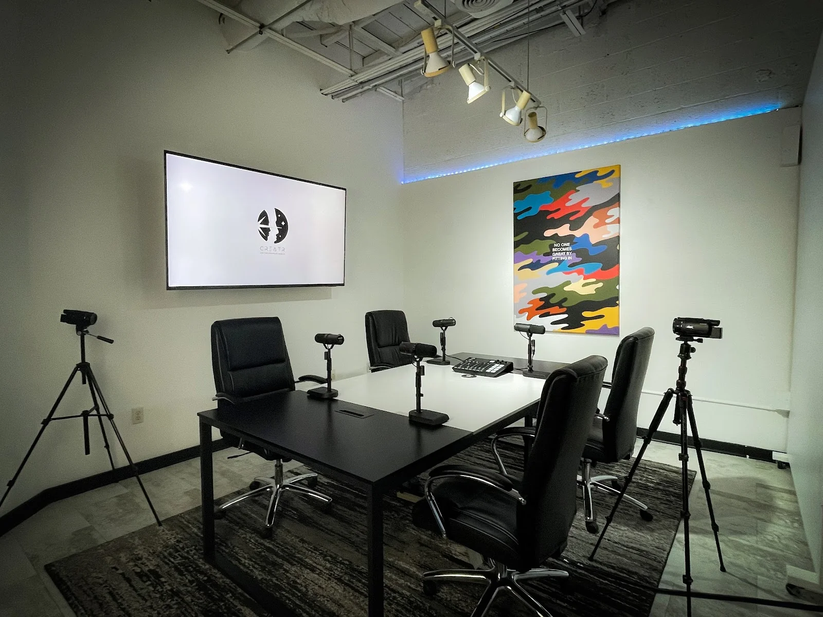 CREATRVERSE podcast and media studio with black conference table, black executive chairs, multiple cameras on tripods, wall-mounted screen showing Creatr logo, colorful camo-print motivational poster, track lighting, and blue LED accent strip along ceiling