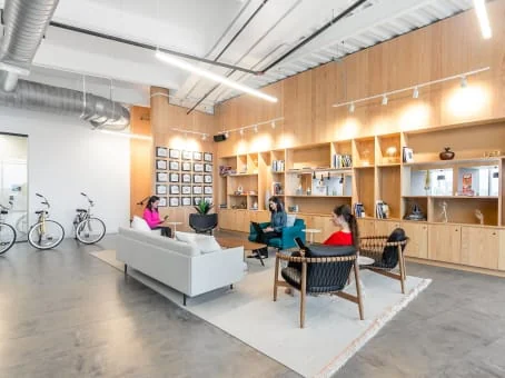 Spaces Deep Ellum modern lounge with warm wood built-in shelving unit displaying books and decor, white sofa, teal accent chair, woven black lounge chair, three women chatting, bicycles parked by wall, exposed ductwork, polished concrete floor, and track lighting
