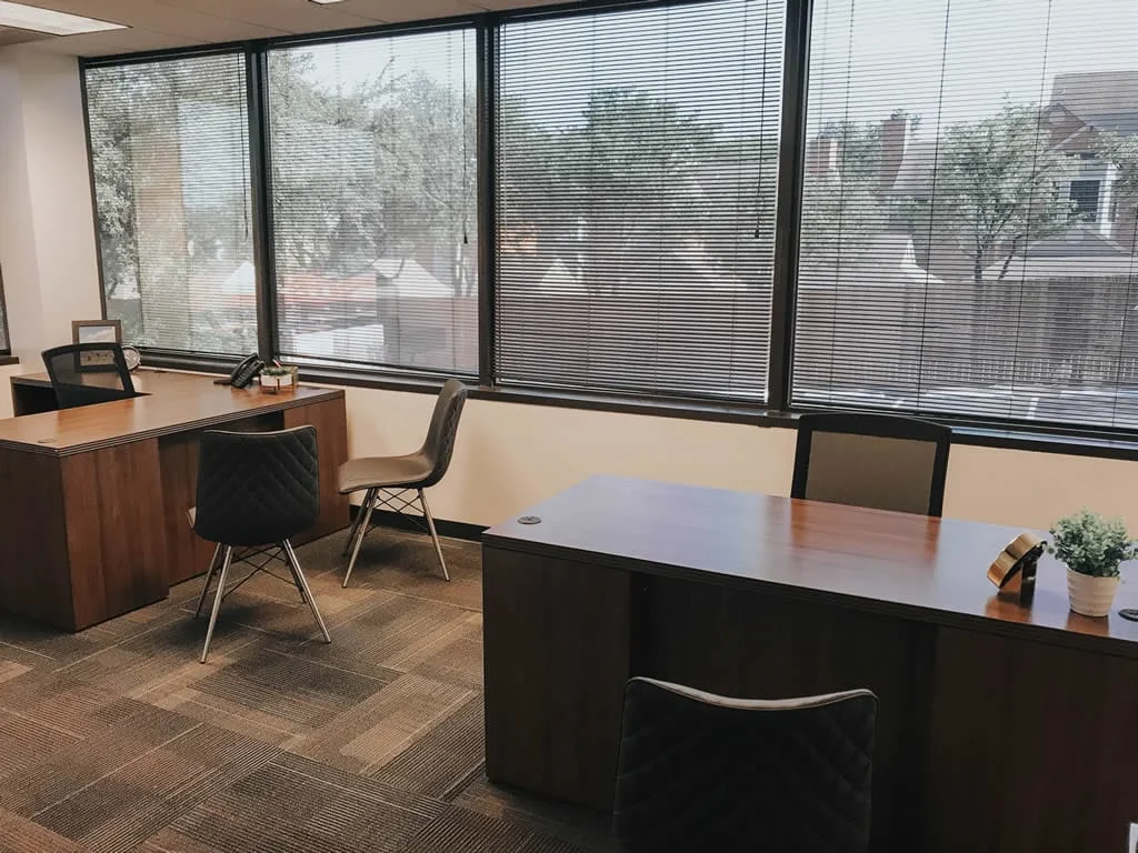ExecutiveWorkspace Hillcrest Green private office with two walnut L-shaped desks, quilted grey and brown accent chairs, large windows with mini-blinds showing tree-lined views, patterned carpet tiles, desk phone, and small plant