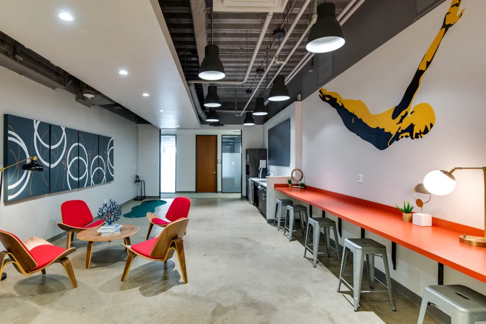 Workstyle Spaces Pacific Ave industrial lounge with red mid-century shell chairs around a small wood table, navy abstract swirl art panels on grey wall, long orange counter bar with metal Tolix stools, bold yellow-and-navy diver wall mural, exposed ceiling with black pendant lights, and polished concrete floors