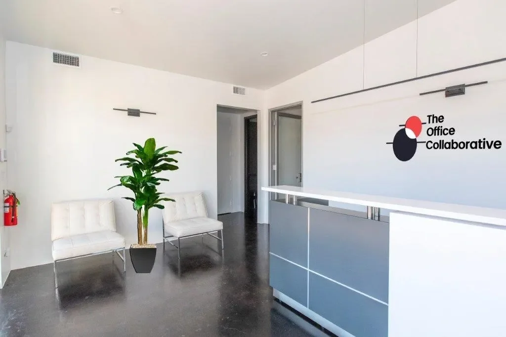 The Office Collaborative minimalist white reception area with red-and-black logo on wall, white Barcelona-style lounge chairs, tropical green plant, sleek grey reception desk with white countertop, dark polished concrete floors, and clean modern picture lights
