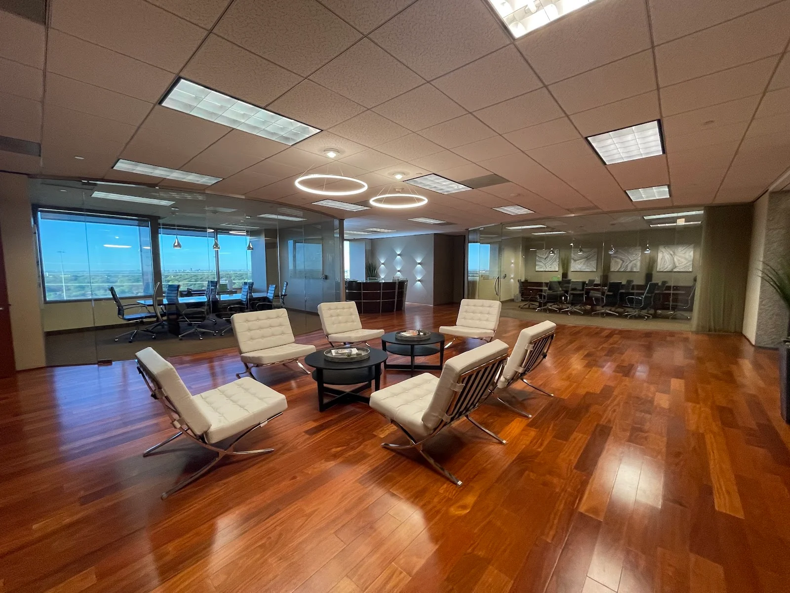 Lucid Private Offices Galleria executive lobby with rich cherry hardwood floors, white Barcelona chairs arranged around a black coffee table, glass-walled conference rooms on both sides, ring pendant lights, and floor-to-ceiling windows with panoramic views