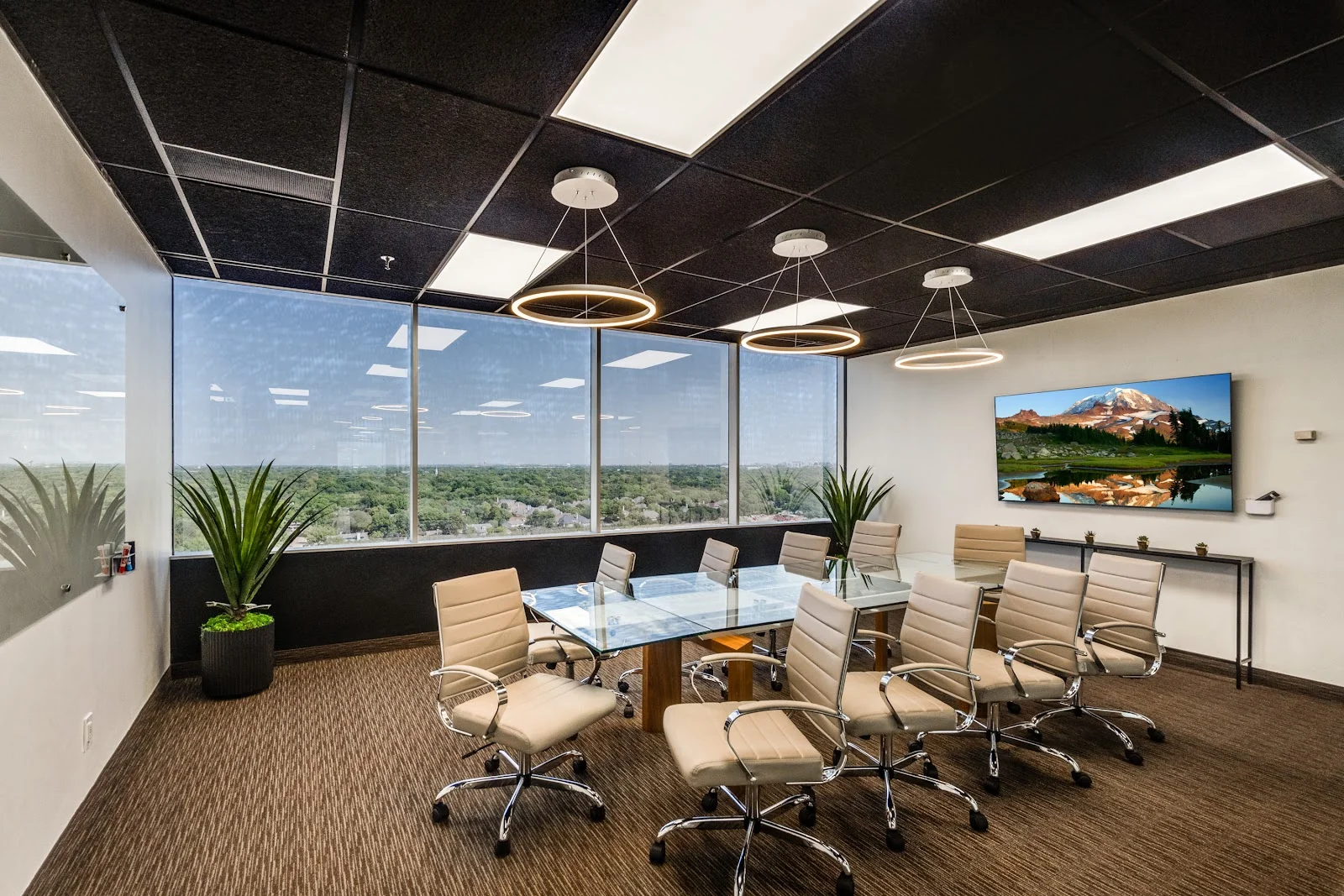 Lucid Private Offices LBJ Freeway high-floor conference room with panoramic window views of suburban Dallas, glass-top table with cream leather executive chairs, black ceiling with ring pendant lights, wall-mounted TV showing mountain landscape, and potted plants