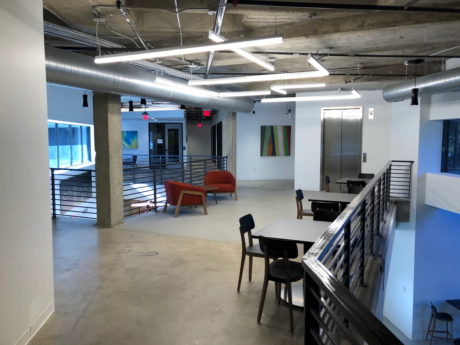 CityCentral North Dallas industrial-modern mezzanine level with polished concrete floors, exposed concrete columns and ceiling, geometric LED light fixtures, metal railings, scattered black chairs and tables, a red accent armchair, and colorful striped art by the elevator