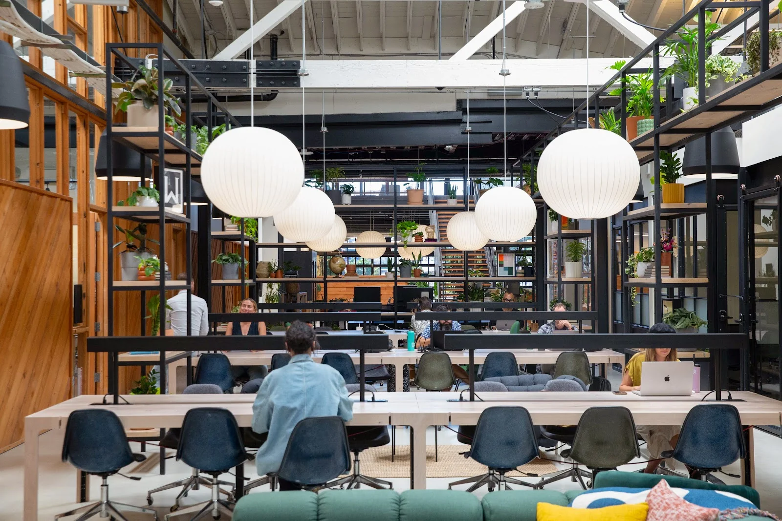 Kiln coworking warehouse with large white globe pendant lights, black steel shelving filled with plants, wooden communal tables, Eames-style chairs, and members working