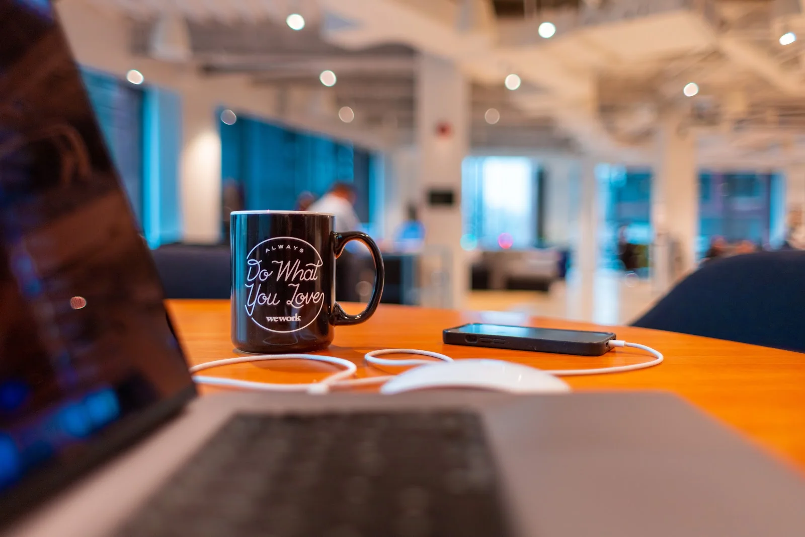 WeWork Do What You Love branded black mug next to laptop and phone on wooden desk in coworking space